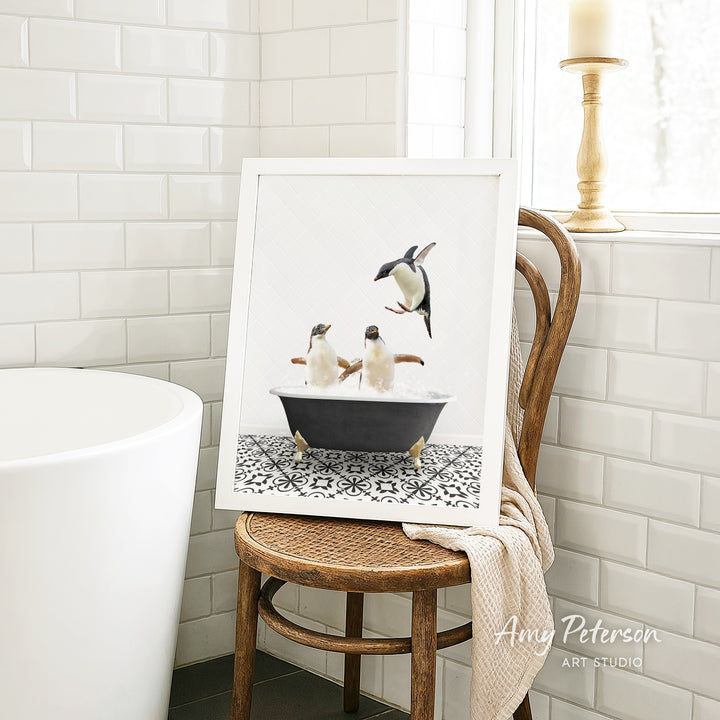 A framed image of two penguins sitting in a bathtub, surrounded by a black and white patterned tile floor, is displayed on a wooden chair in a bathroom setting.