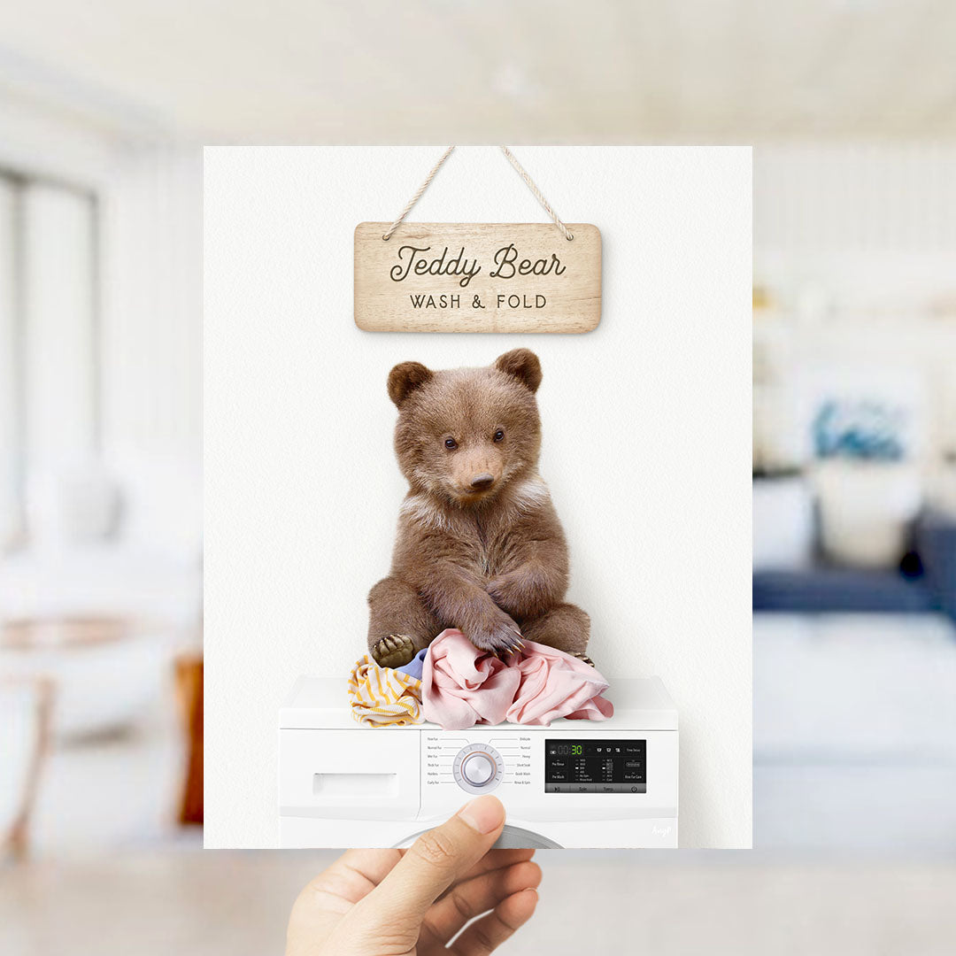 A teddy bear sitting on a pile of clothes inside a washing machine, with a wooden sign above it that says "Teddy Bear WASH & FOLD".