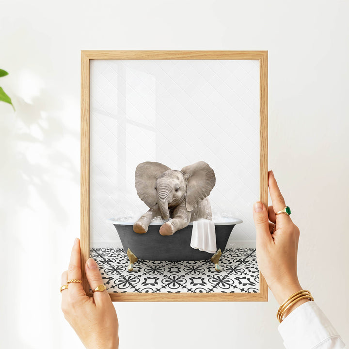 A person's hands are holding a picture frame containing an image of a baby elephant sitting in a bathtub.