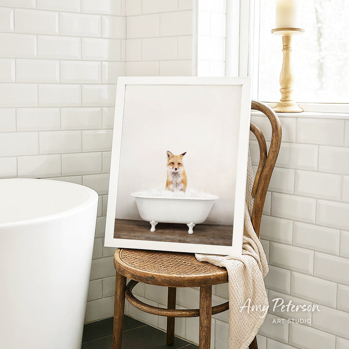 A framed photograph of a fox sitting in a white bathtub, with a towel draped over the back of a wooden chair in a bathroom setting.