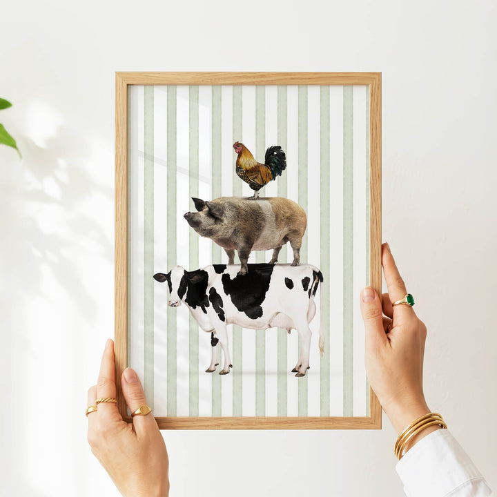 A person's hands are holding a framed picture of a pig, cow, and rooster on a striped background.