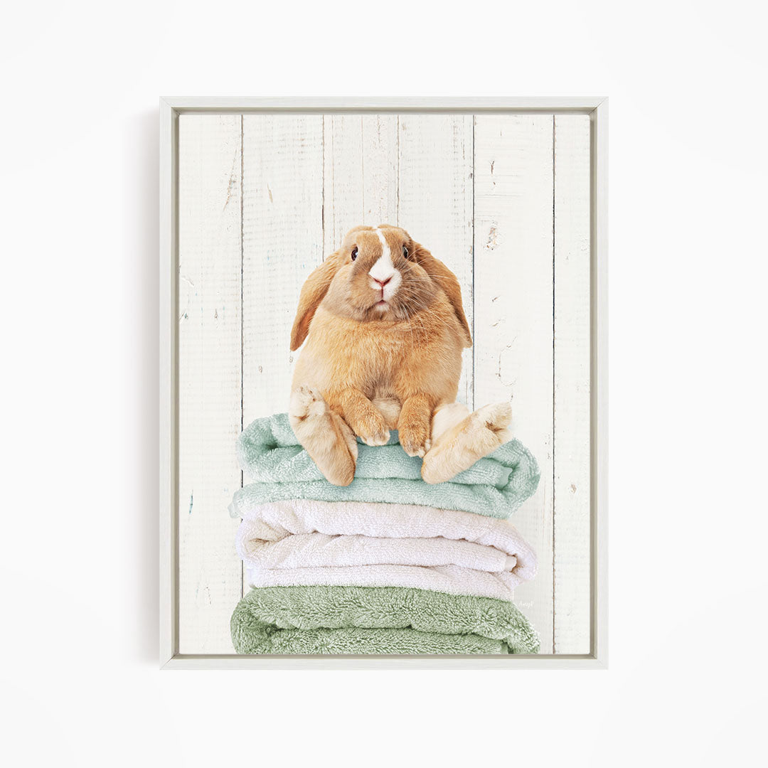 A brown rabbit sitting on a stack of green and white towels against a white wooden background.