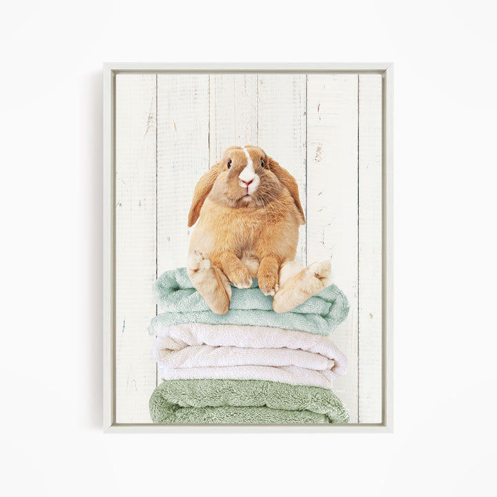 A brown rabbit sitting on a stack of green and white towels against a white wooden background.