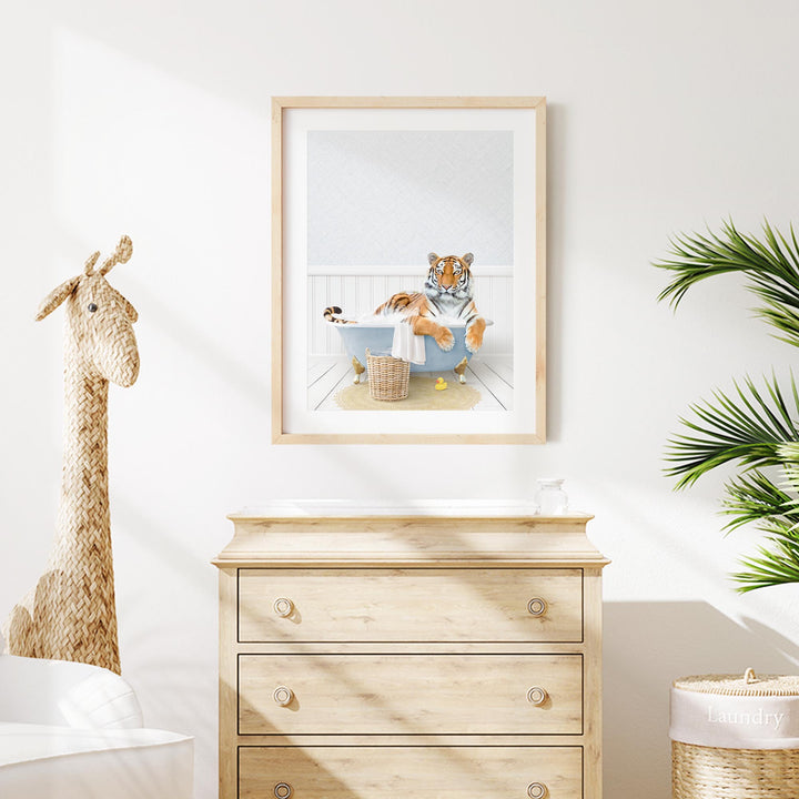 A framed image of a tiger sitting in a bathtub, surrounded by a giraffe and a plant, is displayed on a white wall above a wooden dresser.