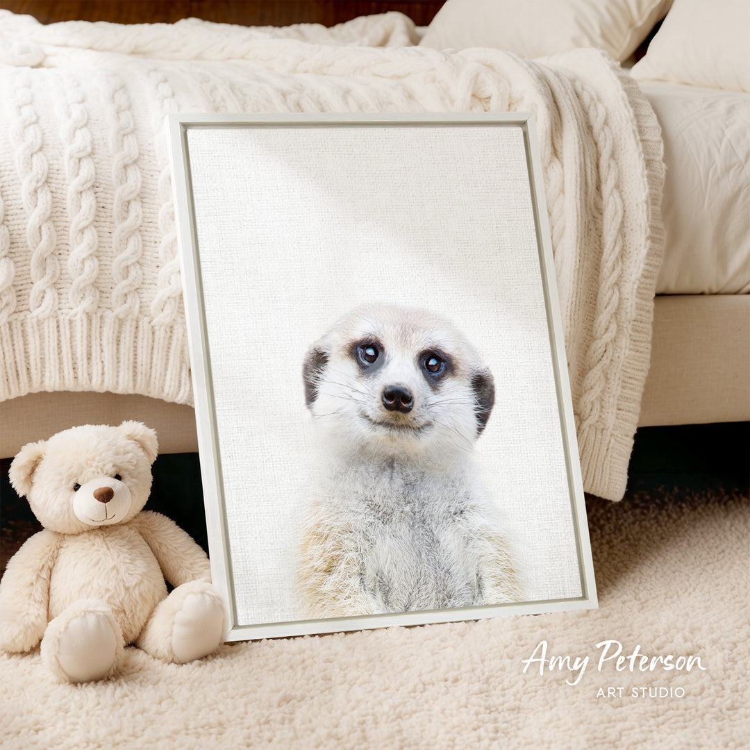 A framed photograph of a meerkat is displayed on a white wall, with a teddy bear sitting next to it on a beige carpet.