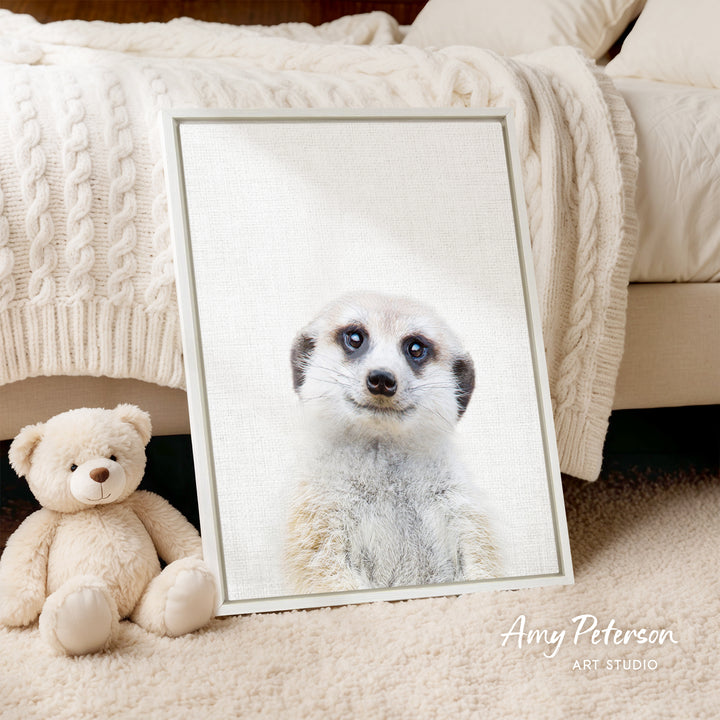 A framed photograph of a meerkat is displayed on a white wall, with a teddy bear sitting next to it on a beige carpet.