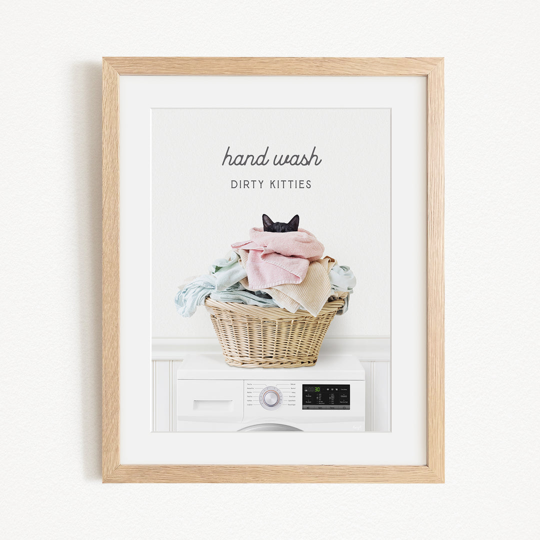 A framed image depicting a cat sitting in a basket filled with laundry, with the text "hand wash dirty kittens" written above the basket.