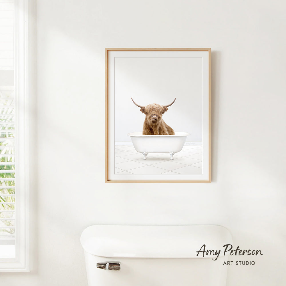 A framed image of a Highland cow sitting in a bathtub is hanging on a white wall.