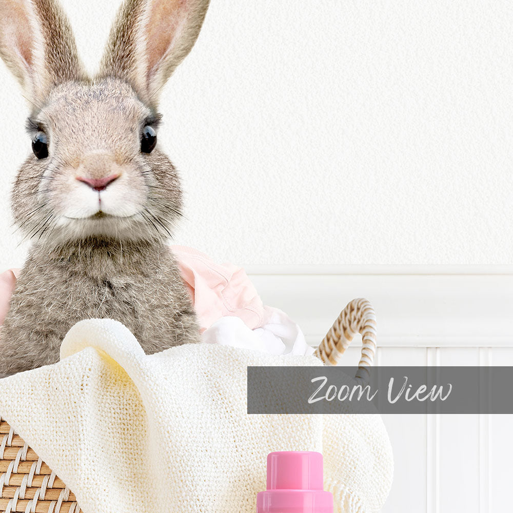A cute gray rabbit peeking out from a basket filled with clothes and a pink bottle.