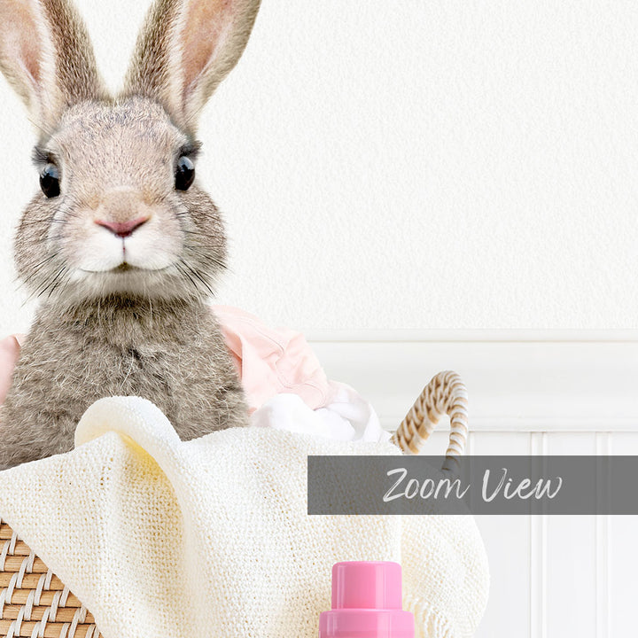 A cute gray rabbit peeking out from a basket filled with clothes and a pink bottle.