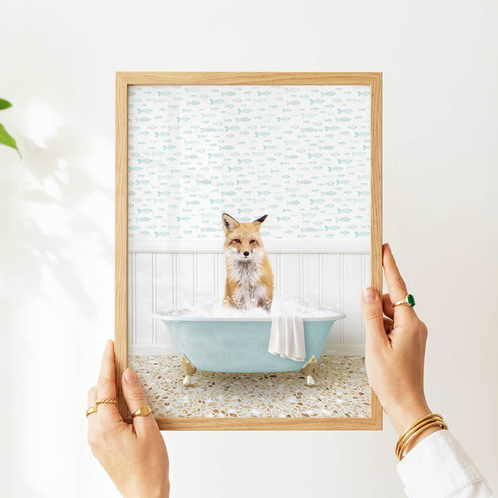 A person's hands are holding a framed picture of a fox sitting in a bathtub filled with bubbles, with a patterned wall behind it.