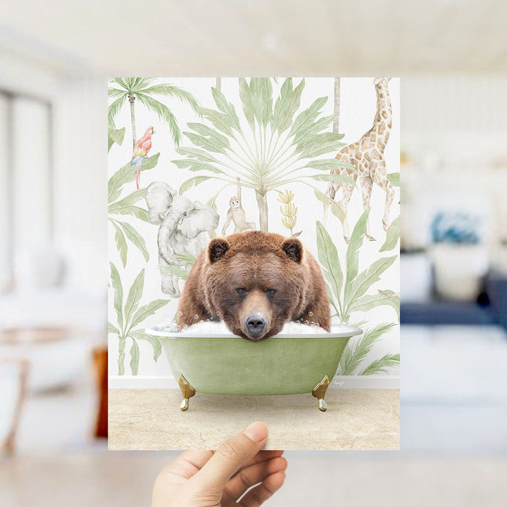 A hand holds up a framed print featuring a brown bear sitting in a green bathtub, surrounded by tropical foliage and animals.