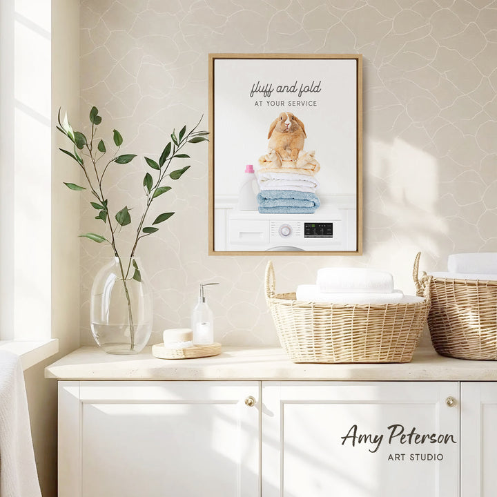 a framed poster of a rabbit sitting on a stack of folded towels, with the text "Fluffy and folded at your service" above it. The poster is displayed on a wall above a white cabinet, which has two woven baskets on top. The cabinet also has a soap dispenser and a plant on the countertop.