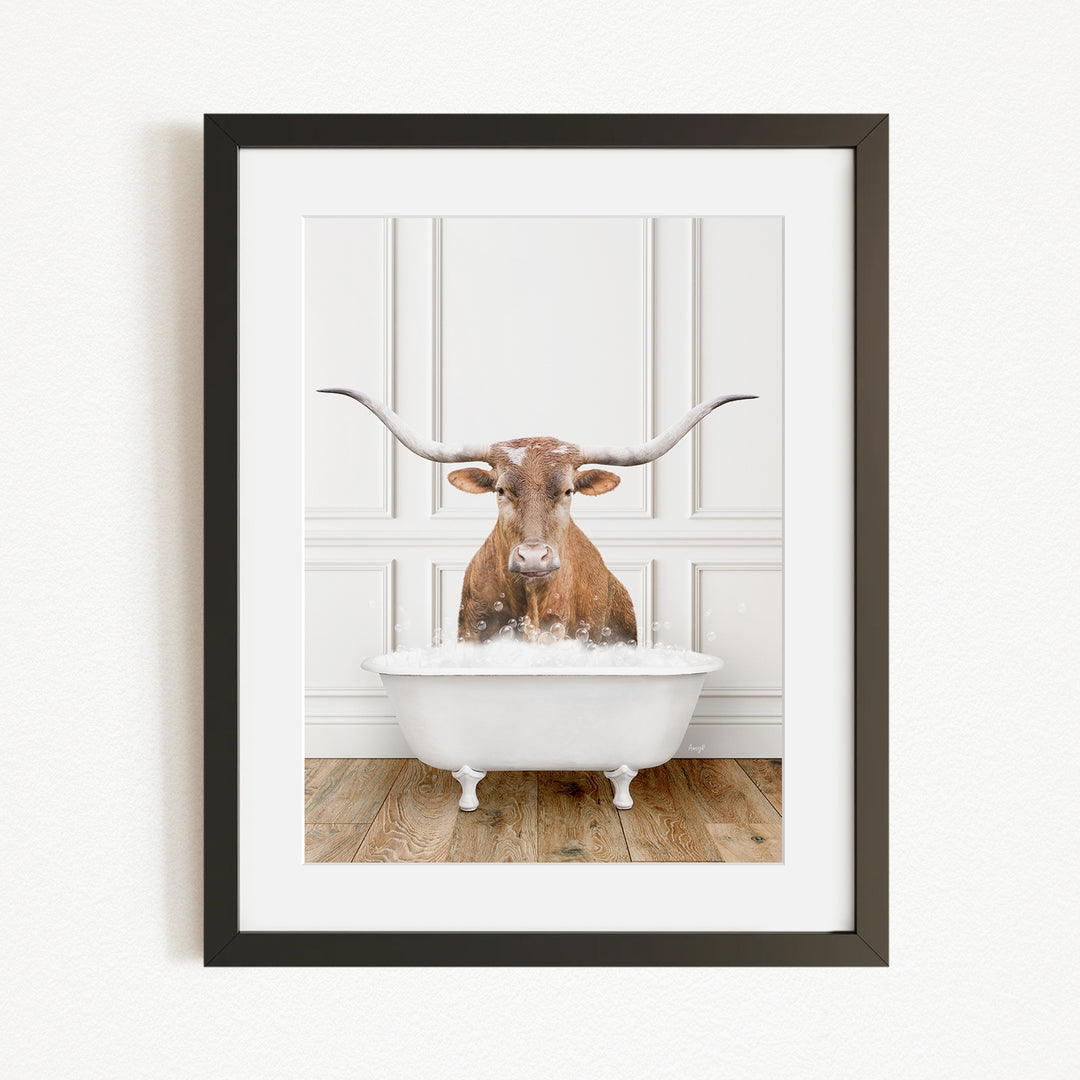 A framed image of a brown cow with long horns sitting in a white bathtub filled with water.