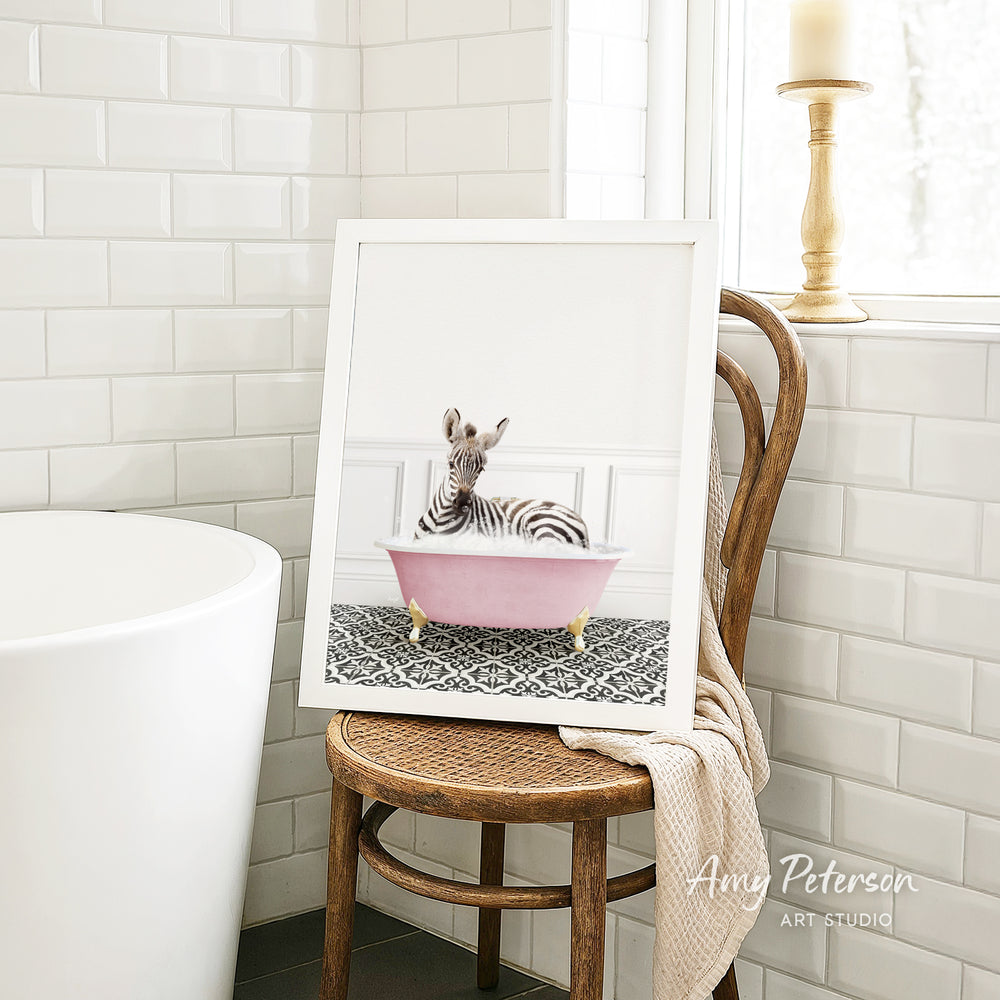 A framed image of a zebra sitting in a pink bathtub is displayed on a wooden chair in a bathroom setting.