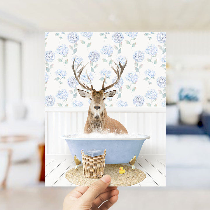 A deer with antlers is sitting in a bathtub filled with bubbles, surrounded by a wicker basket and a rubber duck.