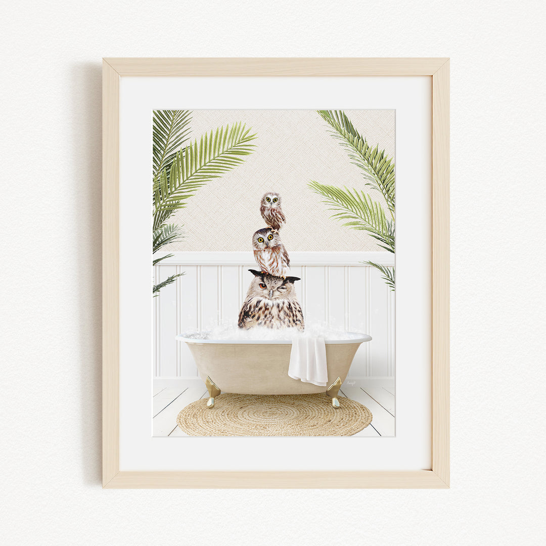 A framed image depicting a white bathtub filled with bubbles, surrounded by lush green palm leaves and a towel, with a pair of owls perched on the edge of the tub.