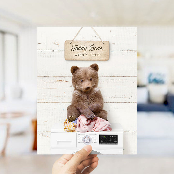 A teddy bear sitting on a pile of clothes inside a washing machine, with a sign above it that says "Teddy Bear Wash & Fold".