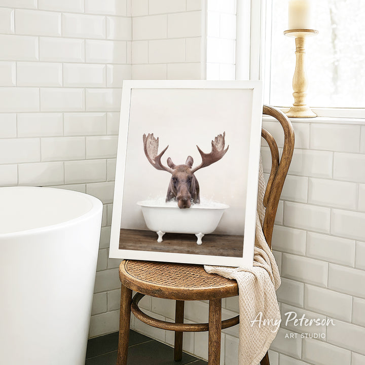 A framed image of a moose with large antlers sitting in a bathtub is displayed on a wooden chair in a bathroom setting.
