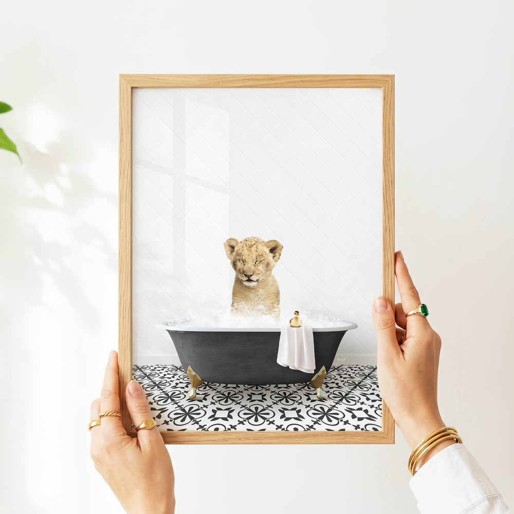 A person's hands are holding a framed picture of a baby lion sitting in a bathtub filled with water, with a towel hanging from the side of the tub.
