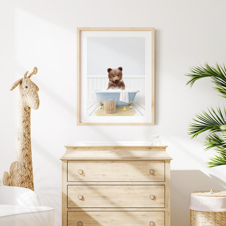 A framed image of a brown bear sitting in a blue bathtub, surrounded by a basket and a towel, is displayed on a white wall above a wooden dresser.