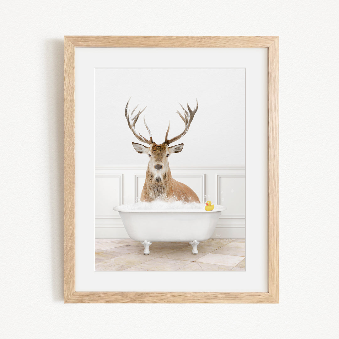 A deer with large antlers is sitting in a white bathtub filled with bubbles, with a yellow rubber duck floating in the water.