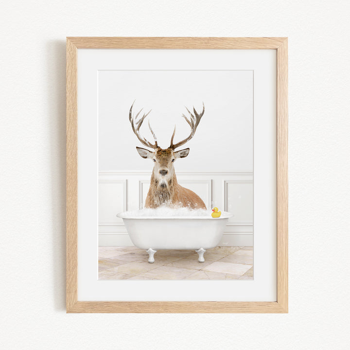 A deer with large antlers is sitting in a white bathtub filled with bubbles, with a yellow rubber duck floating in the water.