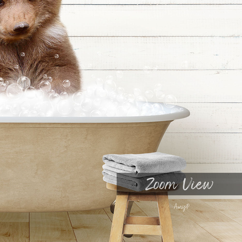 A brown bear is sitting in a bathtub filled with bubbles, with a towel and stool nearby.