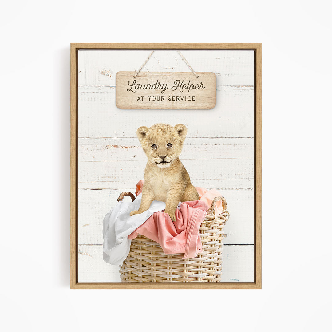 A young lion cub sitting in a wicker basket filled with clothes, with a wooden sign above that reads "Laundry Helper at your service".