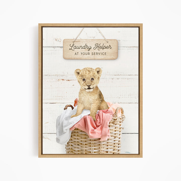 A young lion cub sitting in a wicker basket filled with clothes, with a wooden sign above that reads "Laundry Helper at your service".