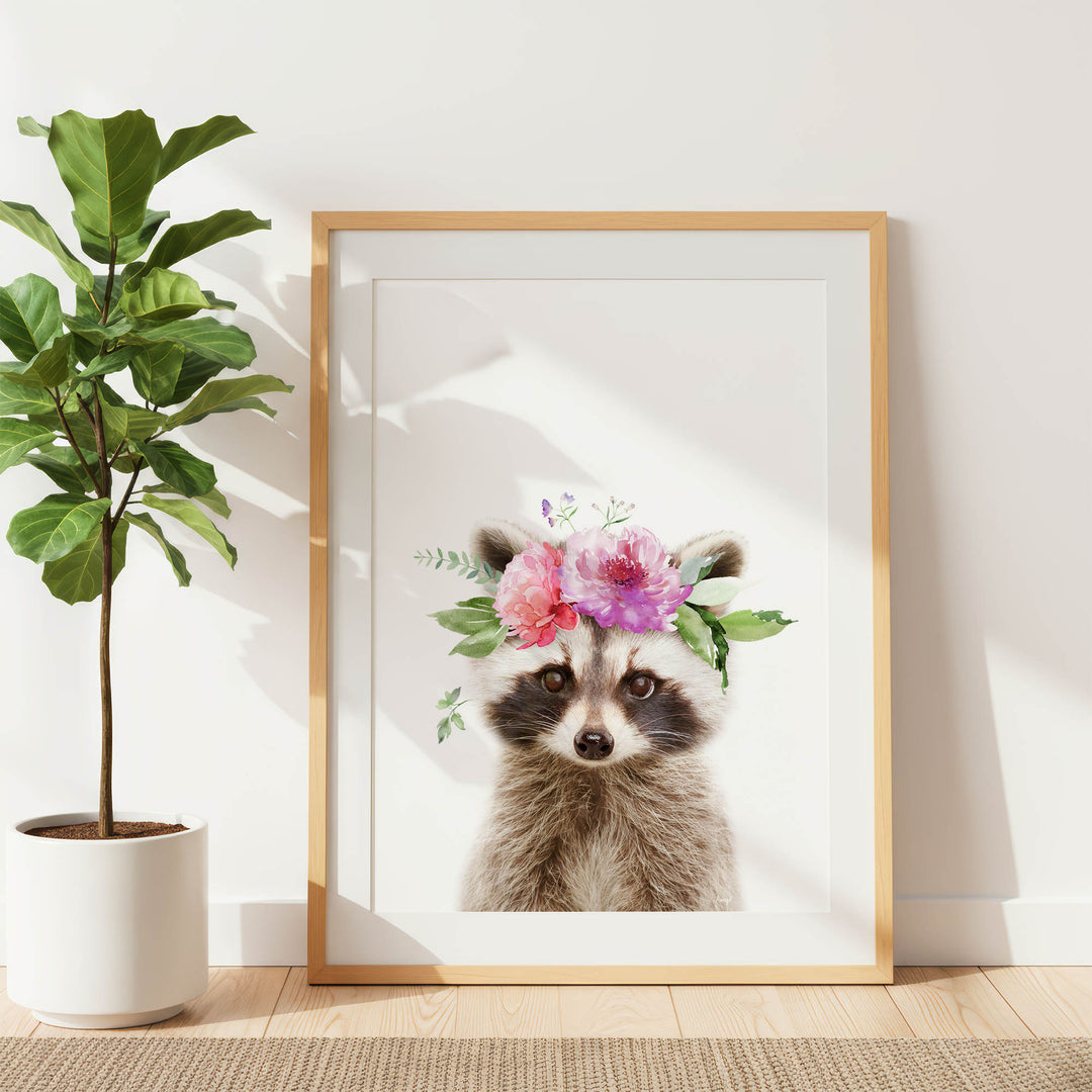 A framed print of a raccoon wearing a floral crown is displayed on a white wall, with a potted plant to the left.
