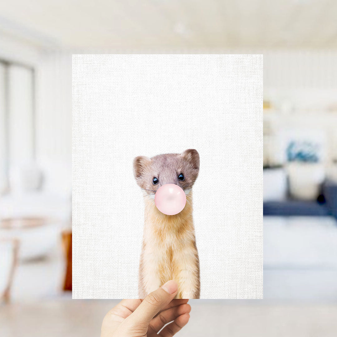 A person's hand is holding up a white square with a small animal, specifically a cat, blowing a bubble with a pink bubble gum bubble.