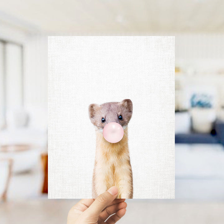 A person's hand is holding up a white square with a small animal, specifically a cat, blowing a bubble with a pink bubble gum bubble.
