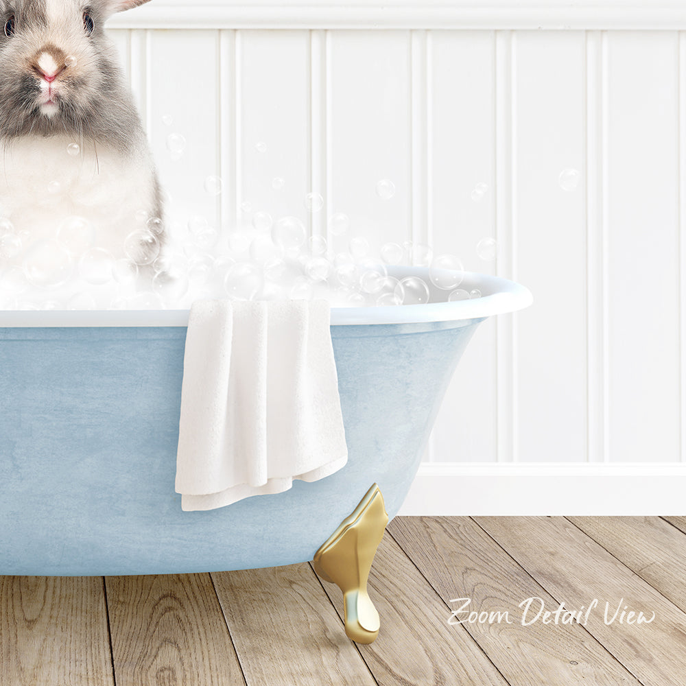 A small rabbit sitting in a blue bathtub filled with bubbles, with a towel and gold bathtub leg visible.