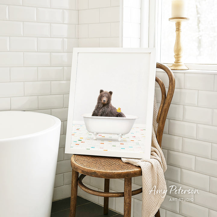 A framed image of a brown bear sitting in a white bathtub, with a yellow rubber duck floating in the water.