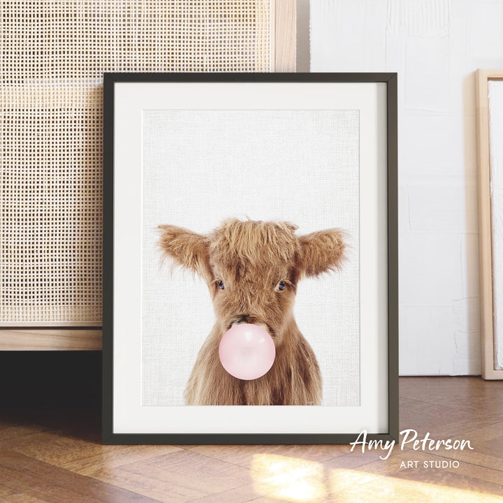 A framed image of a brown cow blowing a bubble with a pink bubble gum bubble.