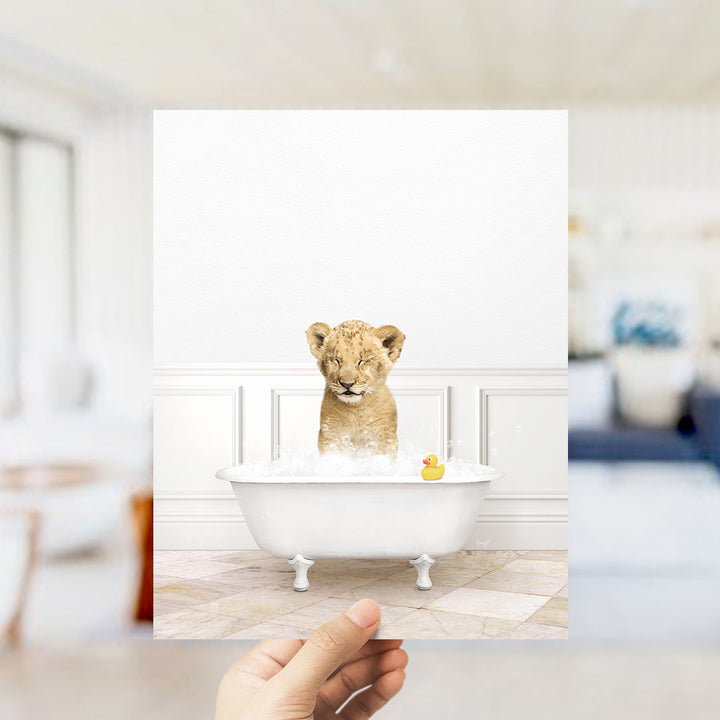 A young lion cub sitting in a white bathtub filled with water and bubbles, with a yellow rubber duck floating nearby.