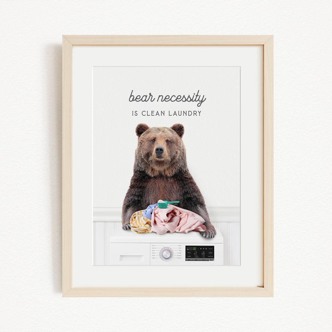 A framed image of a brown bear sitting on a washing machine, with the text "bear necessity is clean laundry" above it.