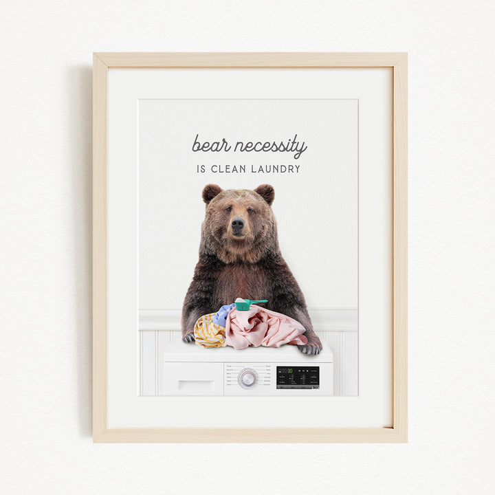 A framed image of a brown bear sitting on a washing machine, with the text "bear necessity is clean laundry" above it.