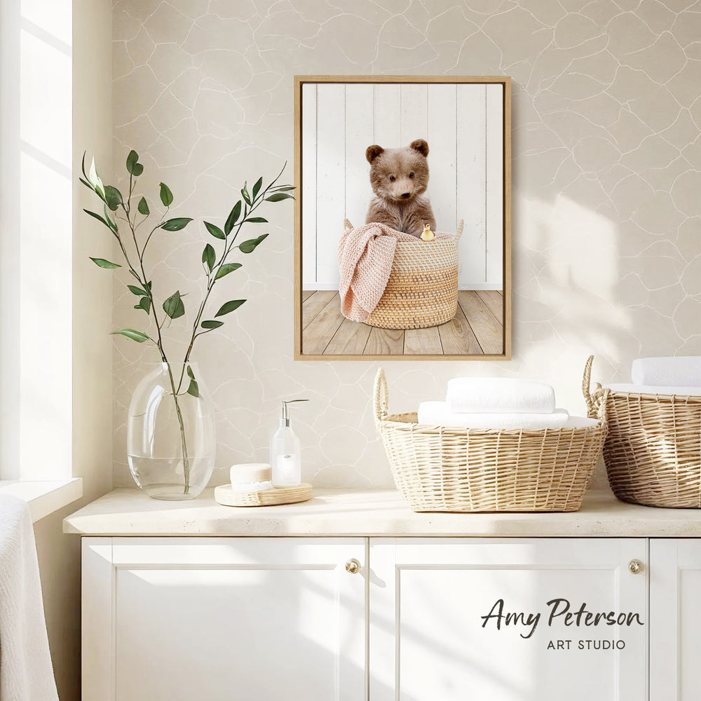 a cozy bathroom scene with a framed photo of a cute bear sitting in a basket, a vase with green leaves, and baskets on a white countertop.