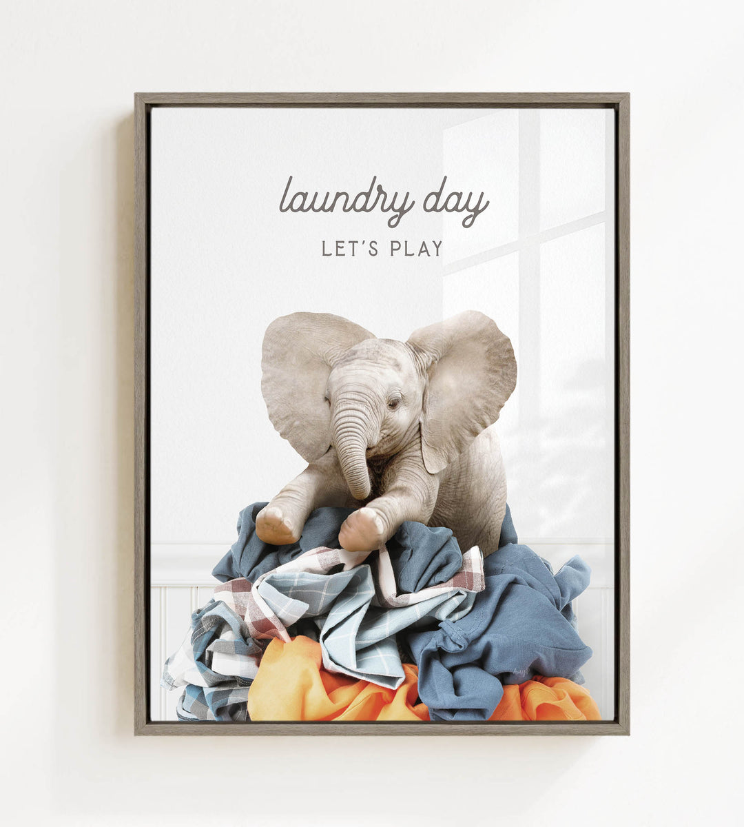 A framed image of a baby elephant sitting on a pile of laundry, with the text "laundry day let's play" written above it.