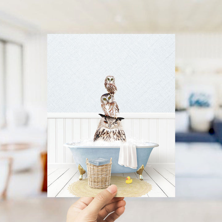 A hand holds up a framed image depicting three owls sitting in a bathtub, with a yellow rubber duck floating nearby.