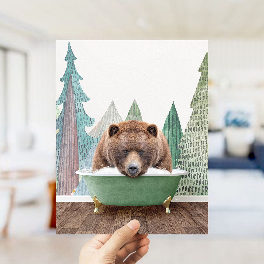 A hand holds up a framed image depicting a brown bear sitting in a green bathtub, surrounded by a forest scene with trees and a blue sky.