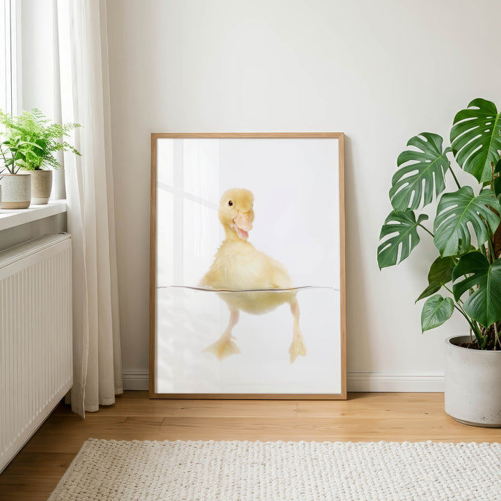 A framed photograph of a yellow duck swimming in a white frame is displayed on a wooden floor in a room with a window and potted plants.