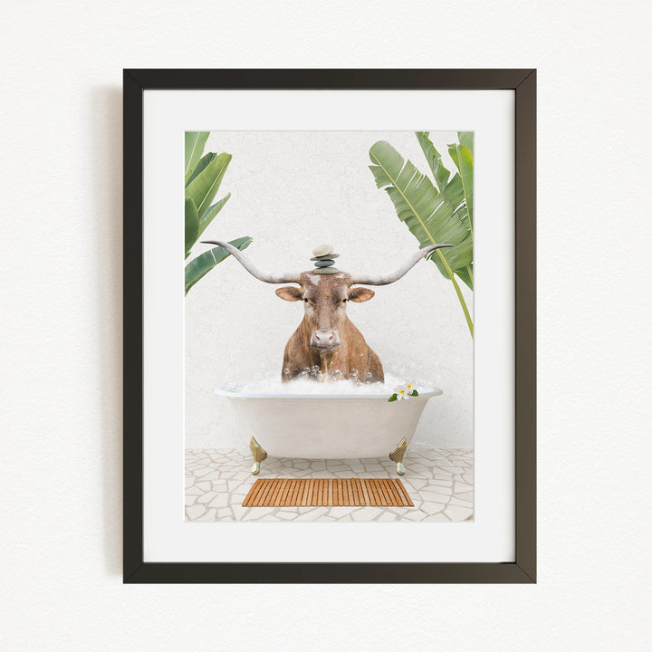 A framed image depicting a cow sitting in a bathtub filled with water, surrounded by palm leaves.