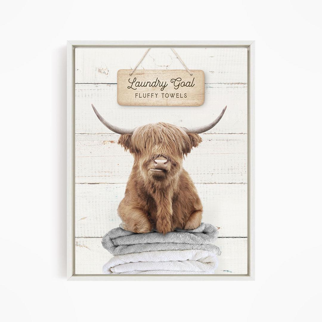 A framed image of a brown cow with long horns sitting on a stack of fluffy towels, with a sign above that says "Laundry Goal Fluffy Towels".