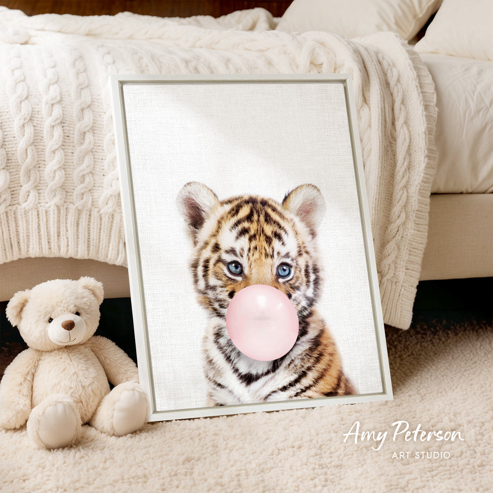 A framed photograph of a tiger cub blowing a bubble with a pink bubble gum bubble, accompanied by a teddy bear on a bed.