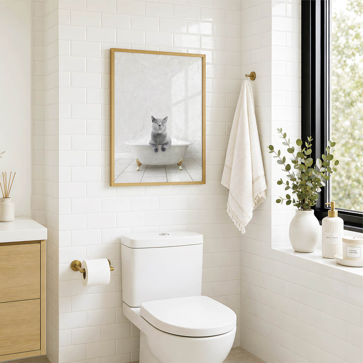 A bathroom with a white toilet, a framed picture of a cat sitting in a bathtub, and a window with greenery on the sill.