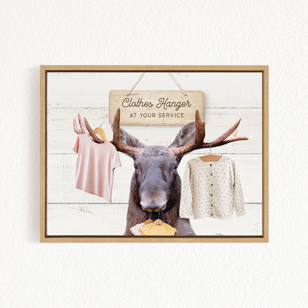 A framed image depicting a moose with antlers, wearing a white sweater and holding a yellow object, with clothes hanging on a clothes hanger above it.