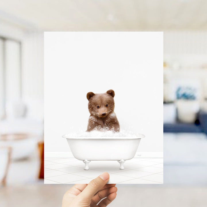A hand holds up a framed image of a brown bear sitting in a white bathtub filled with bubbles.